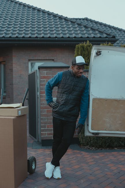 A professional mover from Self Storage Bow stands inside the open rear compartment of a moving van, which is packed with numerous cardboard boxes of various sizes, some sealed with packing tape, and a